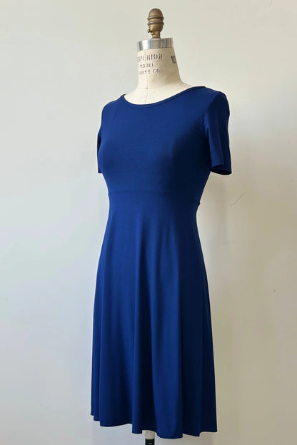 The Luma dress by Kollantai in Admiral blue, featuring short sleeves, a wide neck, an empire waist, and a full flared skirt that falls to the knee, is shown on a mannequin against a white background. 