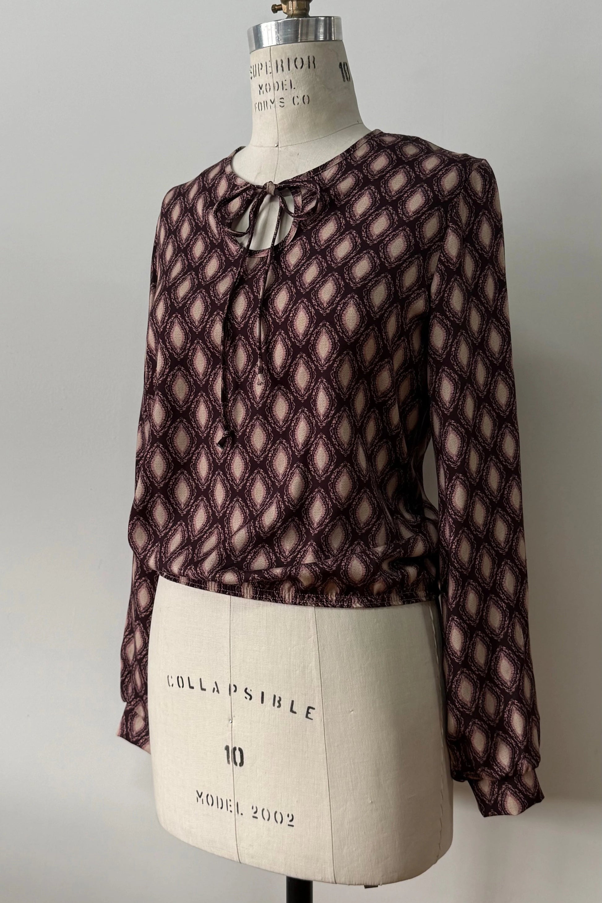 The Isabella Blouse by Kollontai, a burgundy and pink patterned blouse with a tie detail at the neck, balloon sleeves with square jewel buttons, and an elastic waist, is shown on a mannequin against a white background. 