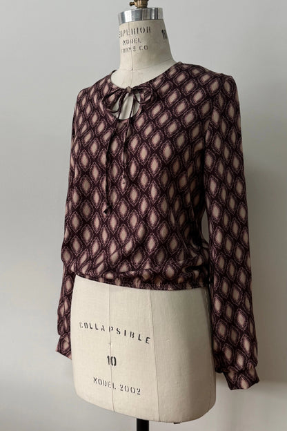 The Isabella Blouse by Kollontai, a burgundy and pink patterned blouse with a tie detail at the neck, balloon sleeves with square jewel buttons, and an elastic waist, is shown on a mannequin against a white background. 