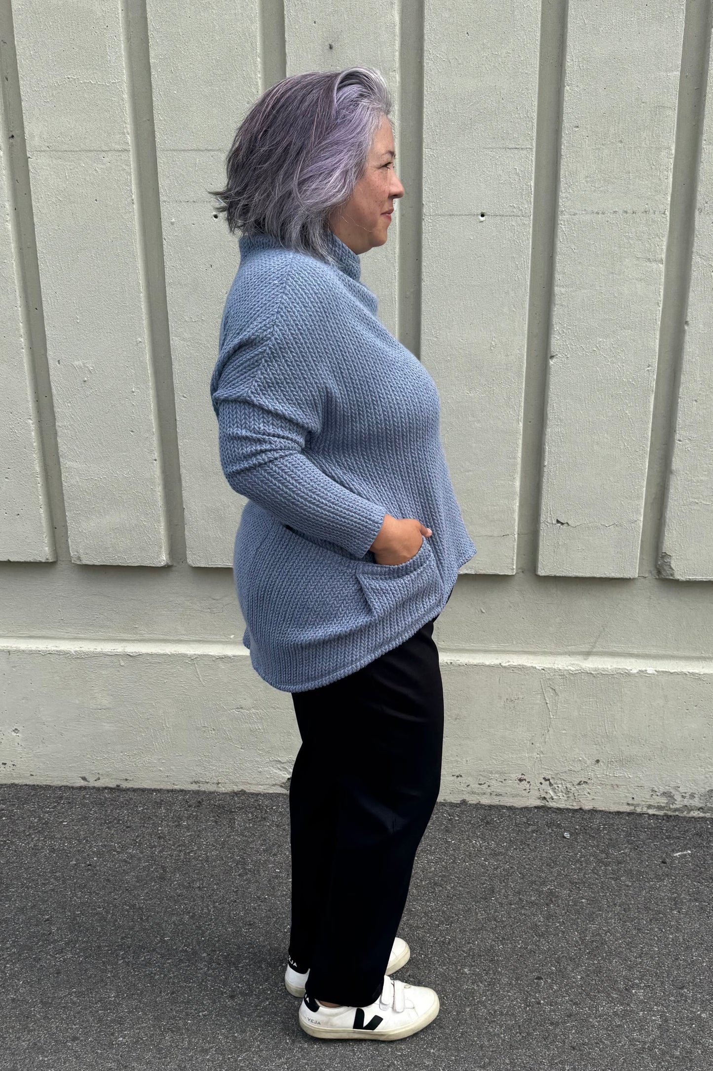 Side view of a woman wearing the Meeso Sweater by MOOVment in Denim, a One Size oversized knit sweater with a wide mock turtleneck, long dropped sleeves and large side pockets. She is wearing it with black pants and standing outside in front of a grey wall. 