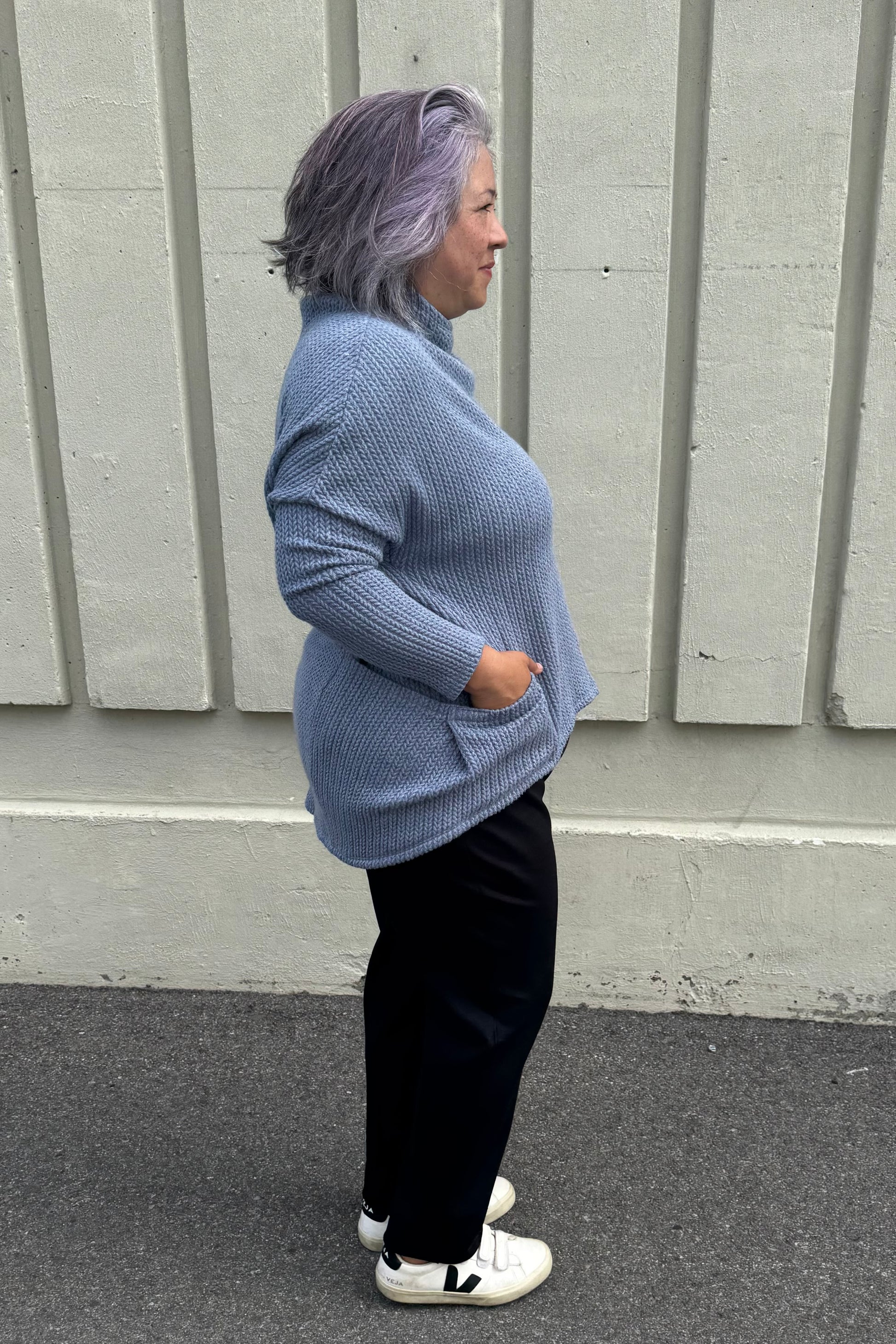 Side view of a woman wearing the Meeso Sweater by MOOVment in Denim, a One Size oversized knit sweater with a wide mock turtleneck, long dropped sleeves and large side pockets. She is wearing it with black pants and standing outside in front of a grey wall. 
