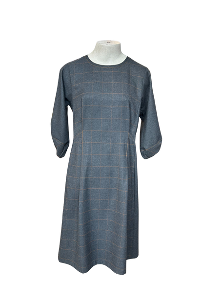 The Hanna Dress by Solomia in Grey is shown on a mannequin against a white background. This knee length dress has uniquely placed pleats and seams to create an architectural shape, with 3/4 length sleeves with structured cuffs. It has checked fabric on the front, back and cuffs, and solid grey fabric at the sides. 