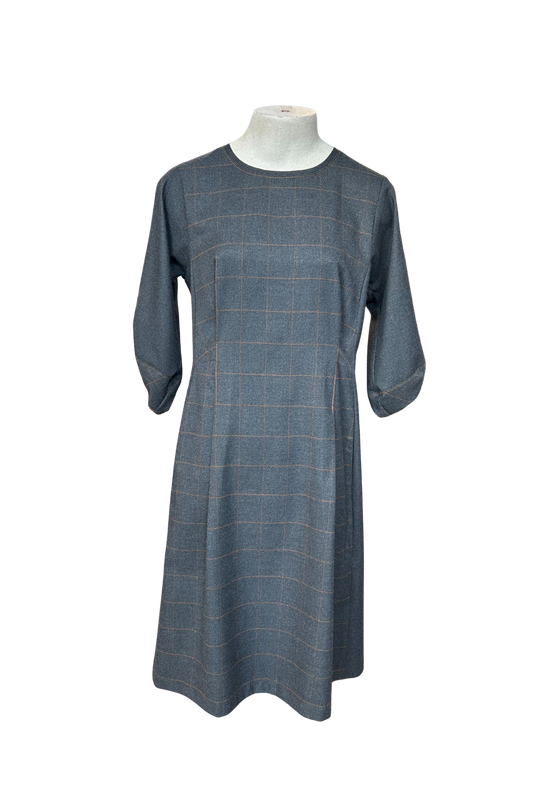 The Hanna Dress by Solomia in Grey is shown on a mannequin against a white background. This knee length dress has uniquely placed pleats and seams to create an architectural shape, with 3/4 length sleeves with structured cuffs. It has checked fabric on the front, back and cuffs, and solid grey fabric at the sides. 