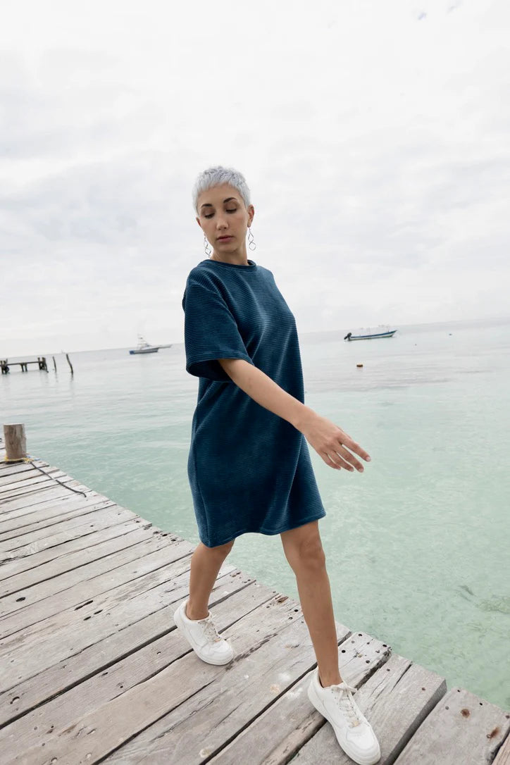 A woman wearing the Mykonos Dress by MAS in Juniper, a short sleeved boxy, knee-length dress in ribbed velvet fabric. She is walking on a wooden dock. 