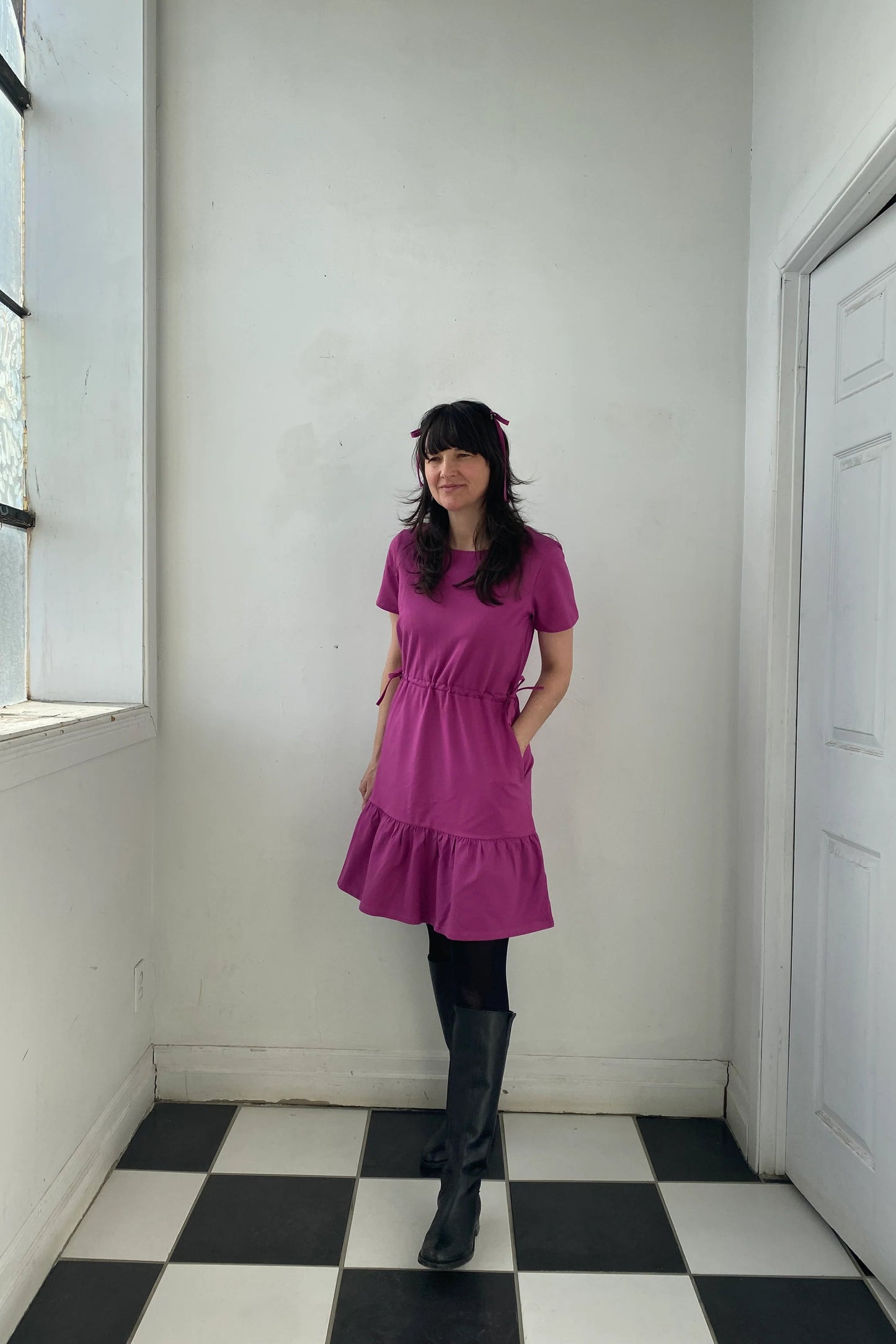 A woman wearing the Épervière Dress by Eve Lavoie in Fuchsia, a reversible dress with short sleeves, a V-neck on one side and a round neck on the other, a flounced hem, and ties at the sides of the waists. She is standing in a room with white walls and black and white checkered floor.