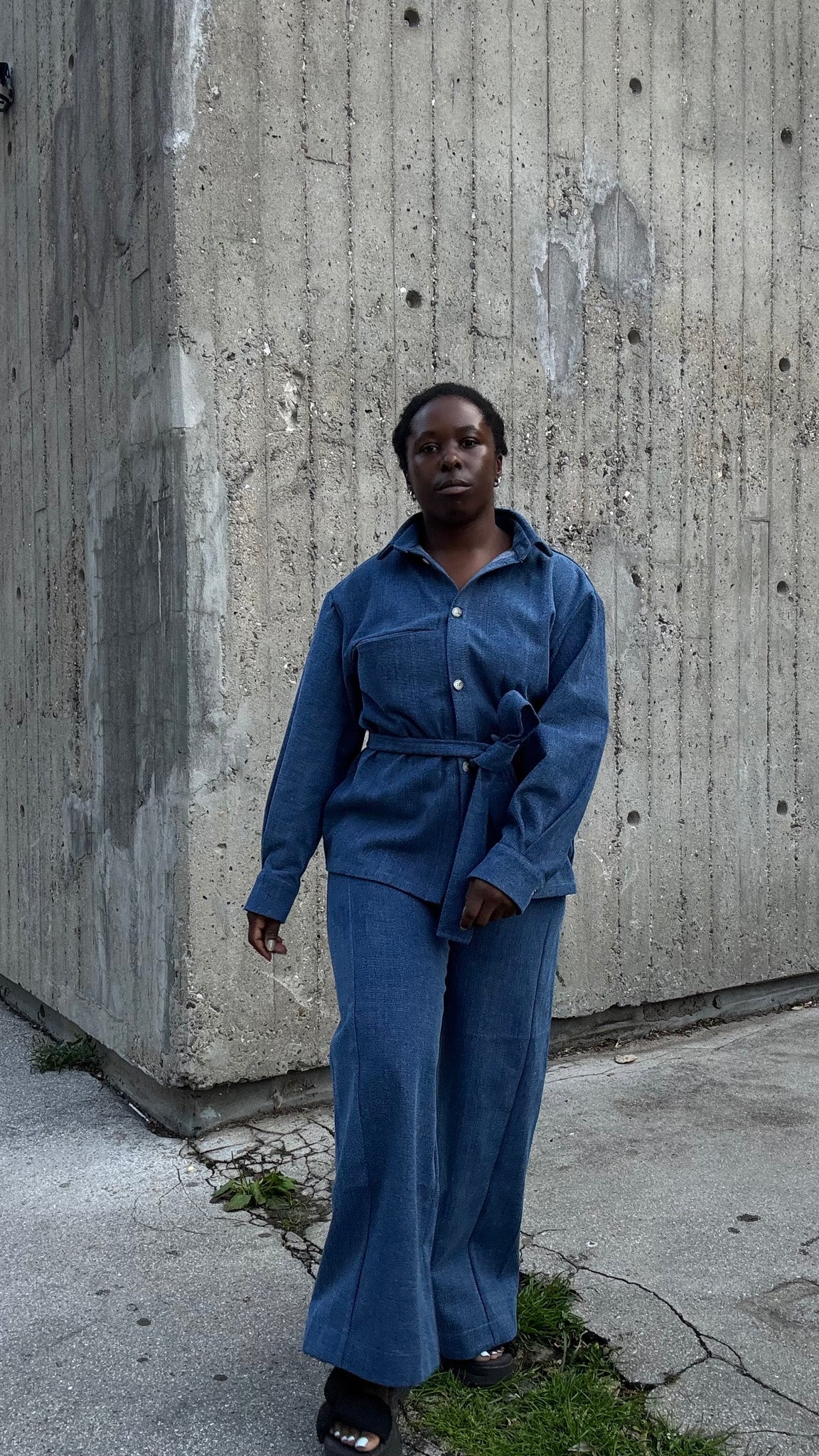  A woman wearing the Vienna Pants by MAS in Denim,  wide legged pants with a high waist and a zip front. She is wearing them with a matching jacket and standing outside by a cement building. 