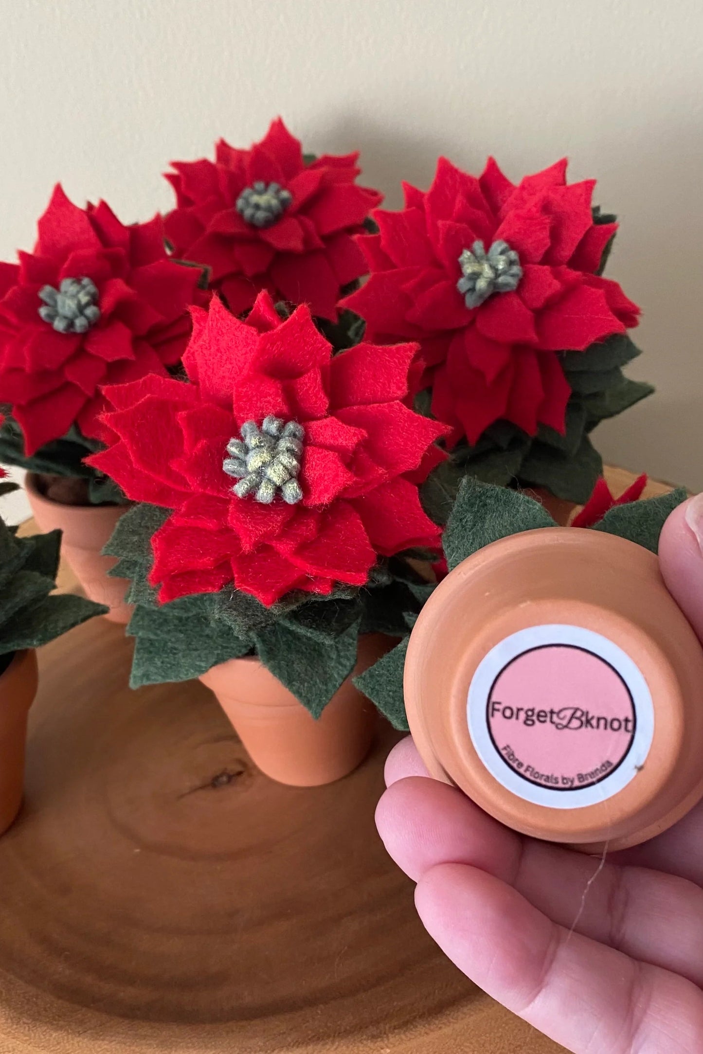 The Holiday Felt Poinsettia by ForgetBKnot is shown on a log against a white background. 