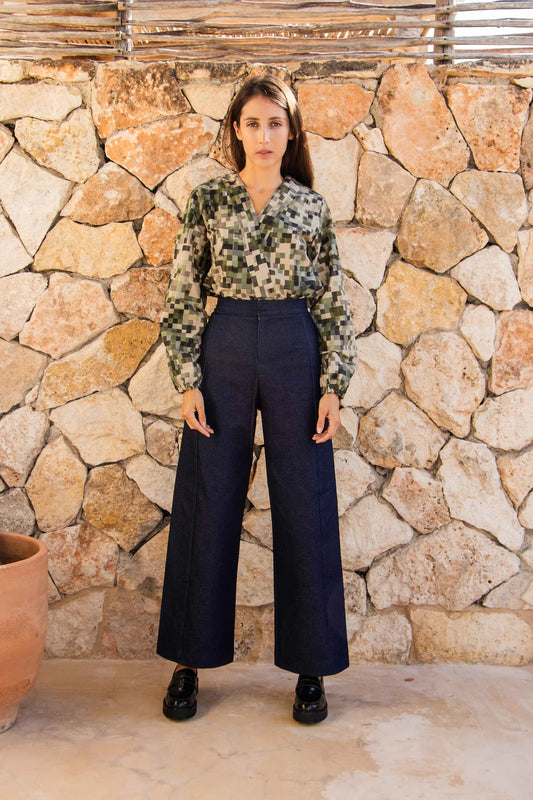 A woman wearing the Vienna Pants by MAS in Jeans, with a wide leg, side seams that sit slightly to the front, and two back pockets, with patterned blouse. She is standing in front of a stone wall. 