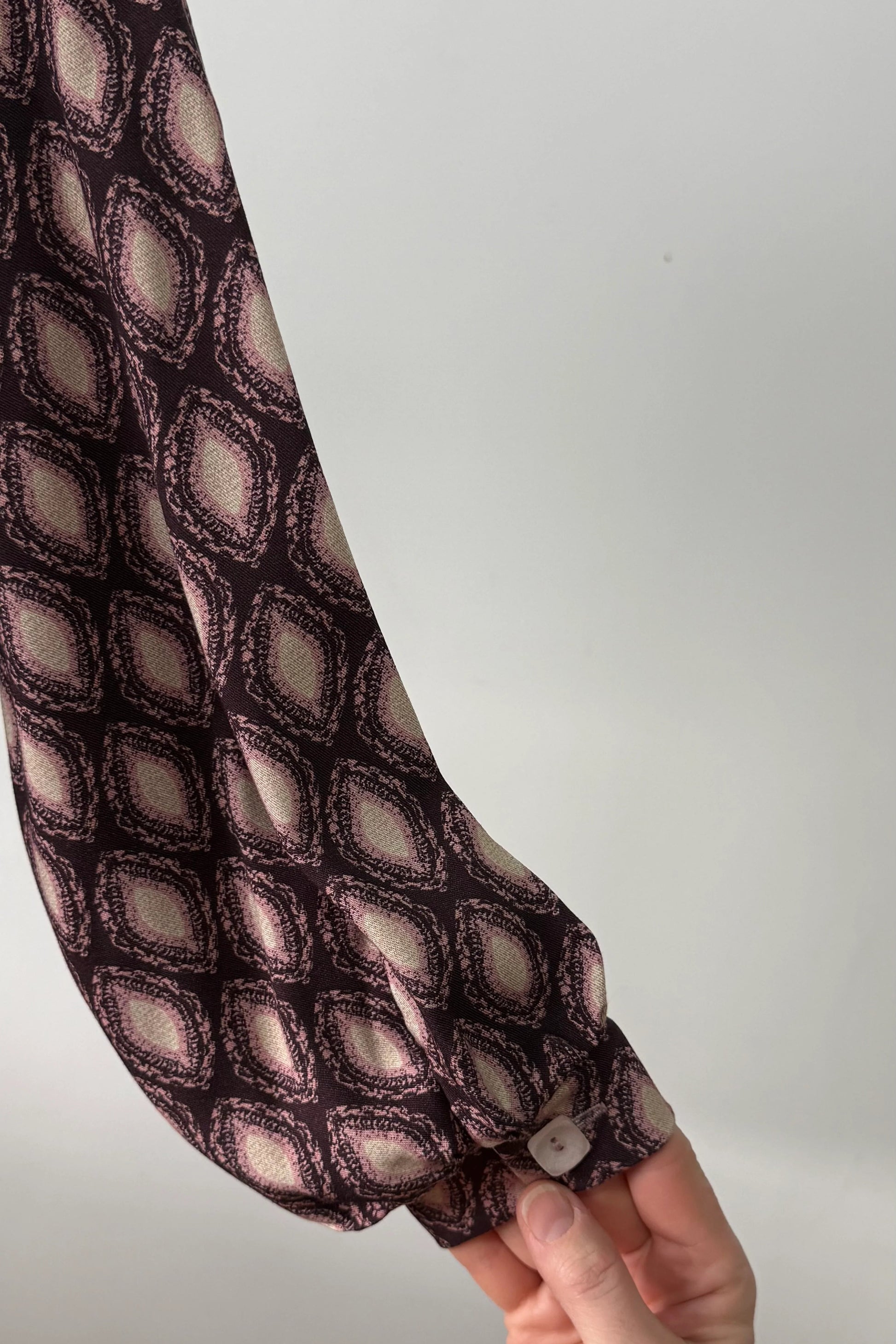 A sleeve detail of the Isabella Blouse by Kollontai, a burgundy and pink patterned blouse with a tie detail at the neck, balloon sleeves with square jewel buttons, and an elastic waist, is shown  against a white background. 