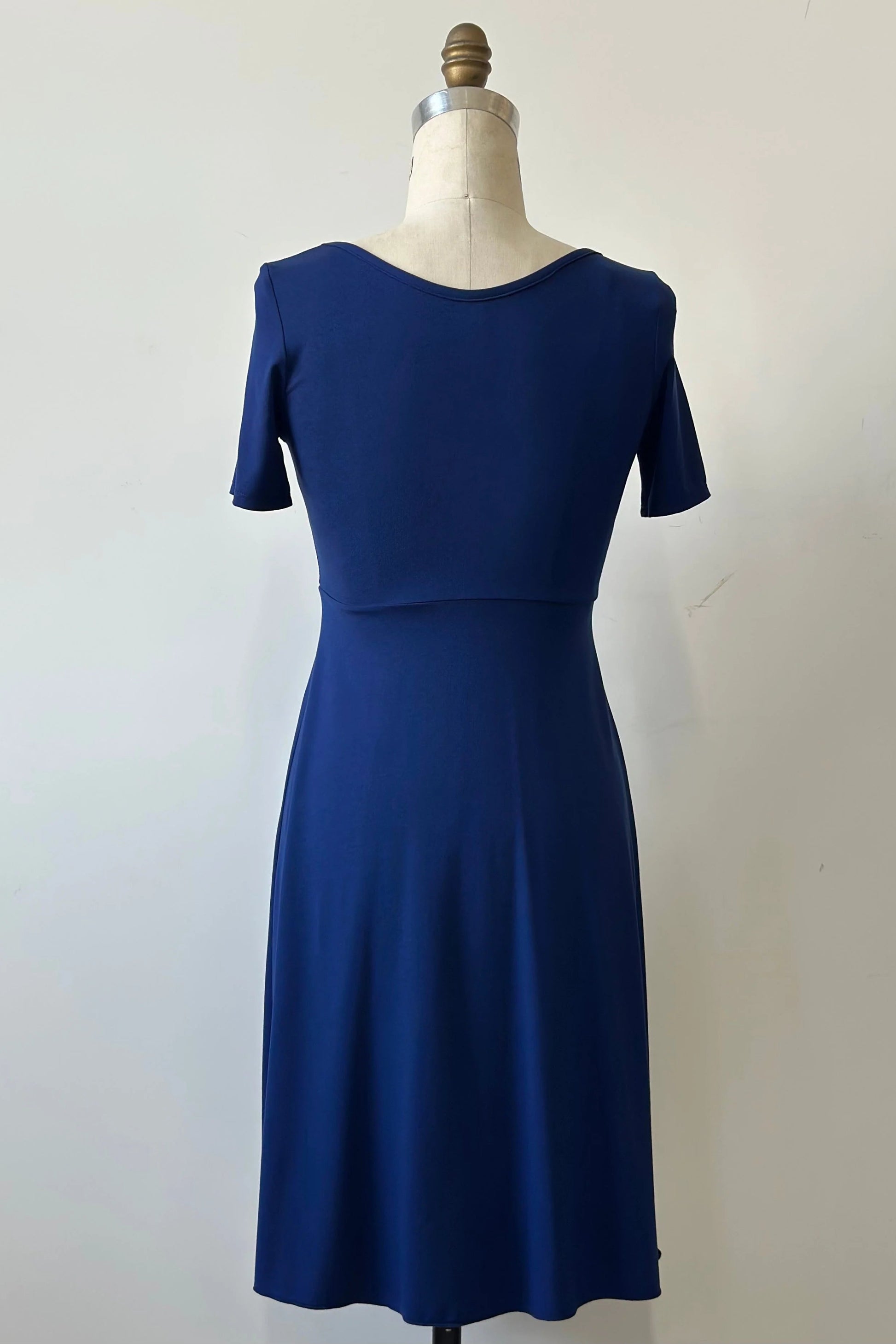 A back view of the Luma dress by Kollantai in Admiral blue, featuring short sleeves, a wide neck, an empire waist, and a full flared skirt that falls to the knee, is shown on a mannequin against a white background. 