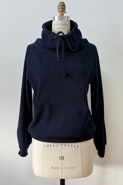 The Ramos Sweater by Kollonta in a ribbed navy corduroy-like fabric, with a large cowl neck with a drawstring and a kangaroo pocket, is shown on a mannequin against a white background. 