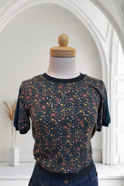 The Darling T-Shirt by Slak in Floral with Green, a short sleeved, semi-fitted sweater with contrasting fabric at the cuffs, collar and hem, inserts on the sleeves, and a solid back, is shown on a mannequin against a white background. 