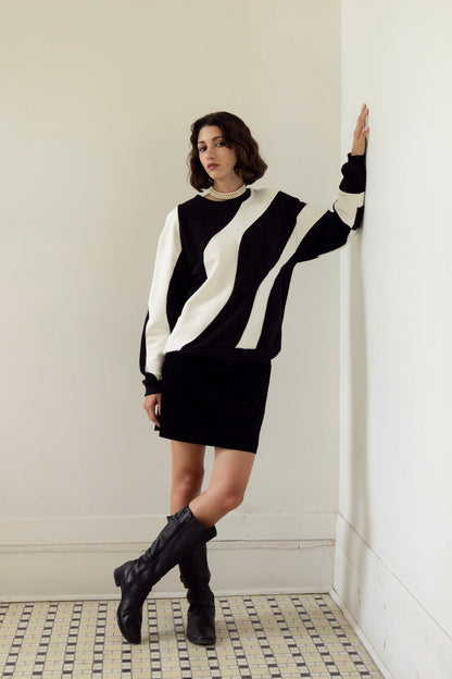 A woman wearing the Amber Sweatshirt by Eve Lavoie in Black/Ivory, an oversized unisex sweatshirt with wave-shaped panels. She is wearing it with a black miniskirt and standing in a white room with a patterned floor.