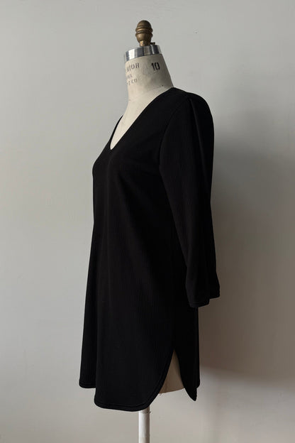 The Clara Tunic by Kollontai in Black, a stretch corduroy piece with a V-neck, a rounded hem, 3/4 sleeves, and inverted pleats at the shoulders, is shown on a mannequin against a grey background. 