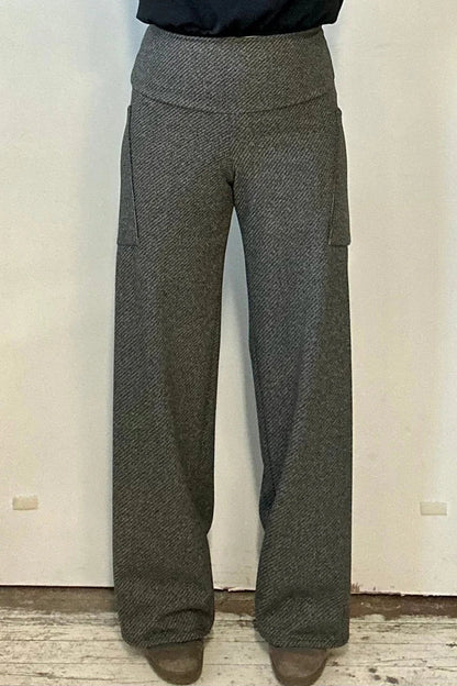 Waist down view of a woman wearing the Susan Pants by Kollontai in Charcoal, wide pants with a wide pull on waistband and angled patch pockets. She is wearing them with a black top and standing in front of a white wall. 