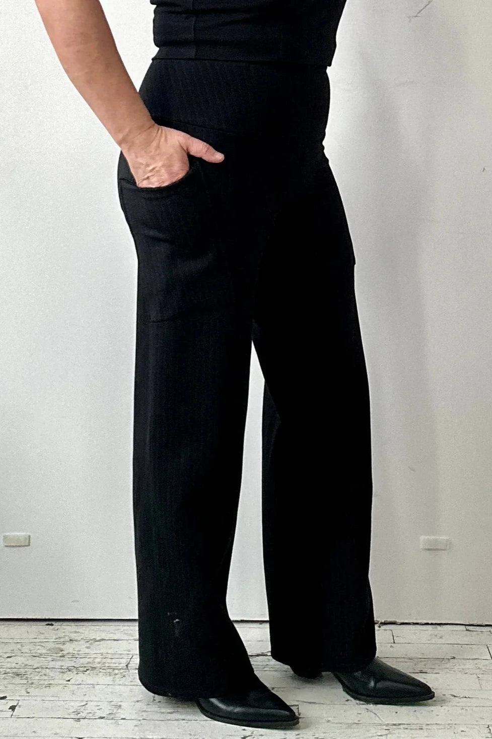 Side waist-down view of a woman wearing the Susan Pants by Kollontai in Black pinstripe, with wide legs, a wide pull-on waistband, and angled side patch pockets. She is standing in front of a white wall. 