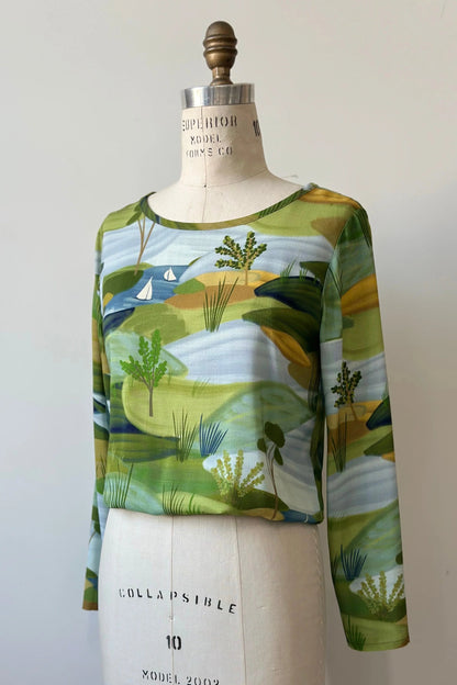 The Belica Blouse by Kollontai in a Granny Smith toned nature print, with a wide neck, long sleeves, linen-look fabric, and an elasticized waist for a subtle blouson effect, is shown on a mannequin in front of a white background. 