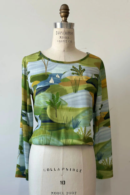 The Belica Blouse by Kollontai in a Granny Smith toned nature print, with a wide neck, long sleeves, linen-look fabric, and an elasticized waist for a subtle blouson effect, is shown on a mannequin in front of a white background. 