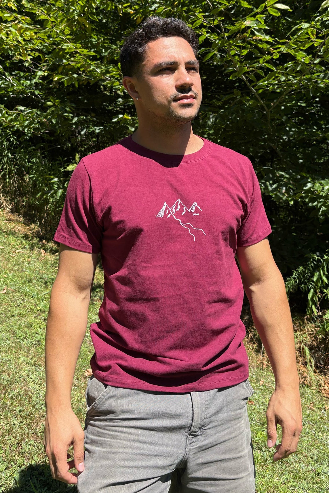 A man wearing the Appalaches T-Shirt by Message Factory, featuring a white mountains graphic on a burgundy background. He is wearing it with grey shorts and standing outside. 