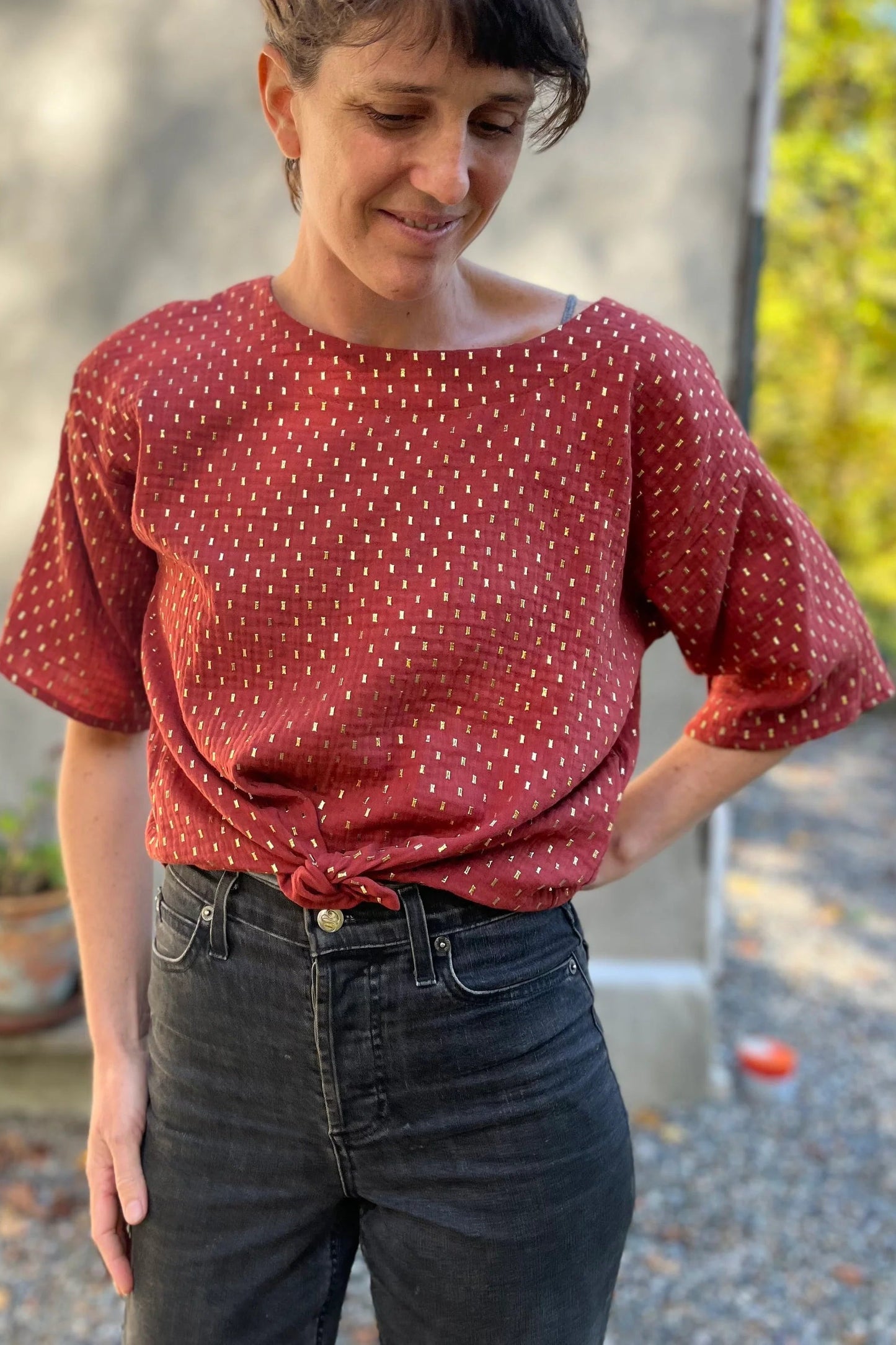 A woman wearing the Abelia Top by Kazak in Golden Tomettes, a boxy shirt with a wide neck and 3/4 kimono sleeves featuring a.red base with gold flecks. She is wearing it with grey jeans and standing outside against a grey wall. 