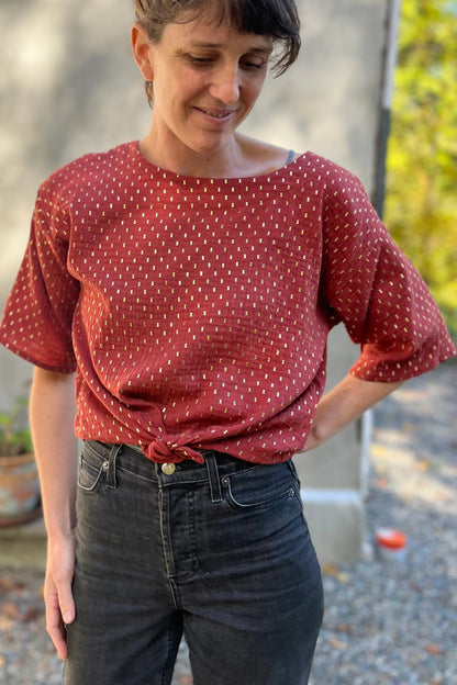 A woman wearing the Abelia Top by Kazak in Golden Tomettes, a boxy shirt with a wide neck and 3/4 kimono sleeves featuring a.red base with gold flecks. She is wearing it with grey jeans and standing outside against a grey wall. 