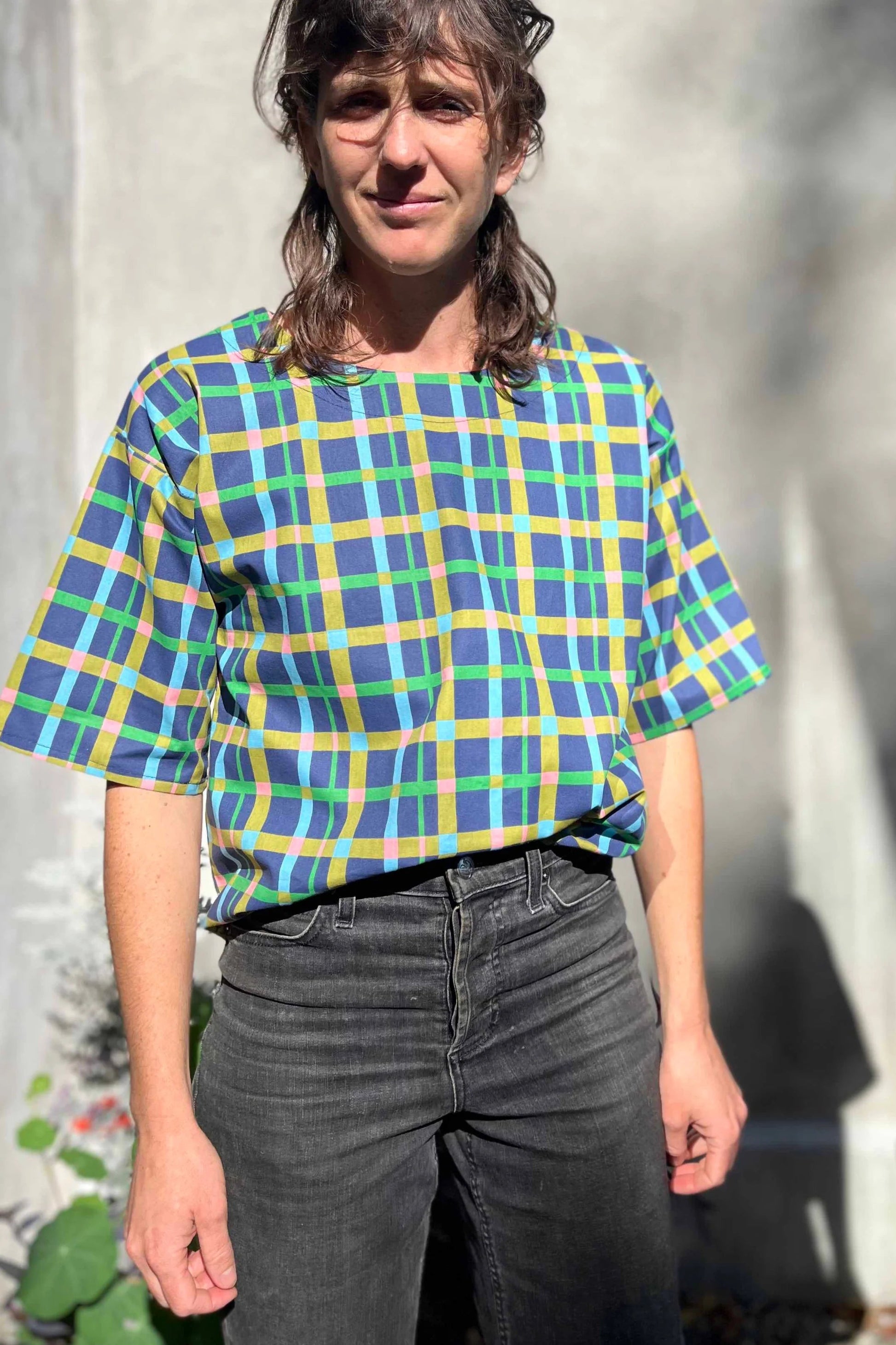 A woman wearing the Abelia Top by Kazak in Blue checks, a boxy shirt with a wide neck and 3/4 kimono sleeves. She is wearing it with grey jeans and standing outside against a grey wall. 
