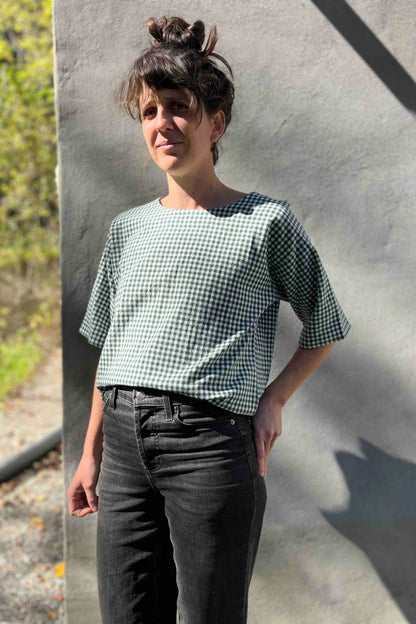A woman wearing the Abelia Top by Kazak in Forest Vichy, a boxy shirt with a wide neck and 3/4 kimono sleeves. She is wearing it with grey jeans and standing outside against a grey wall. 