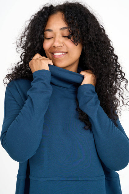 Close up of a woman wearing the Abigail Turtleneck by Advika in Bluebell, a loose fitting turtleneck top with dropped sleeves, a seam across the middle, and a rounded hem. She is standing in front of a white background. 