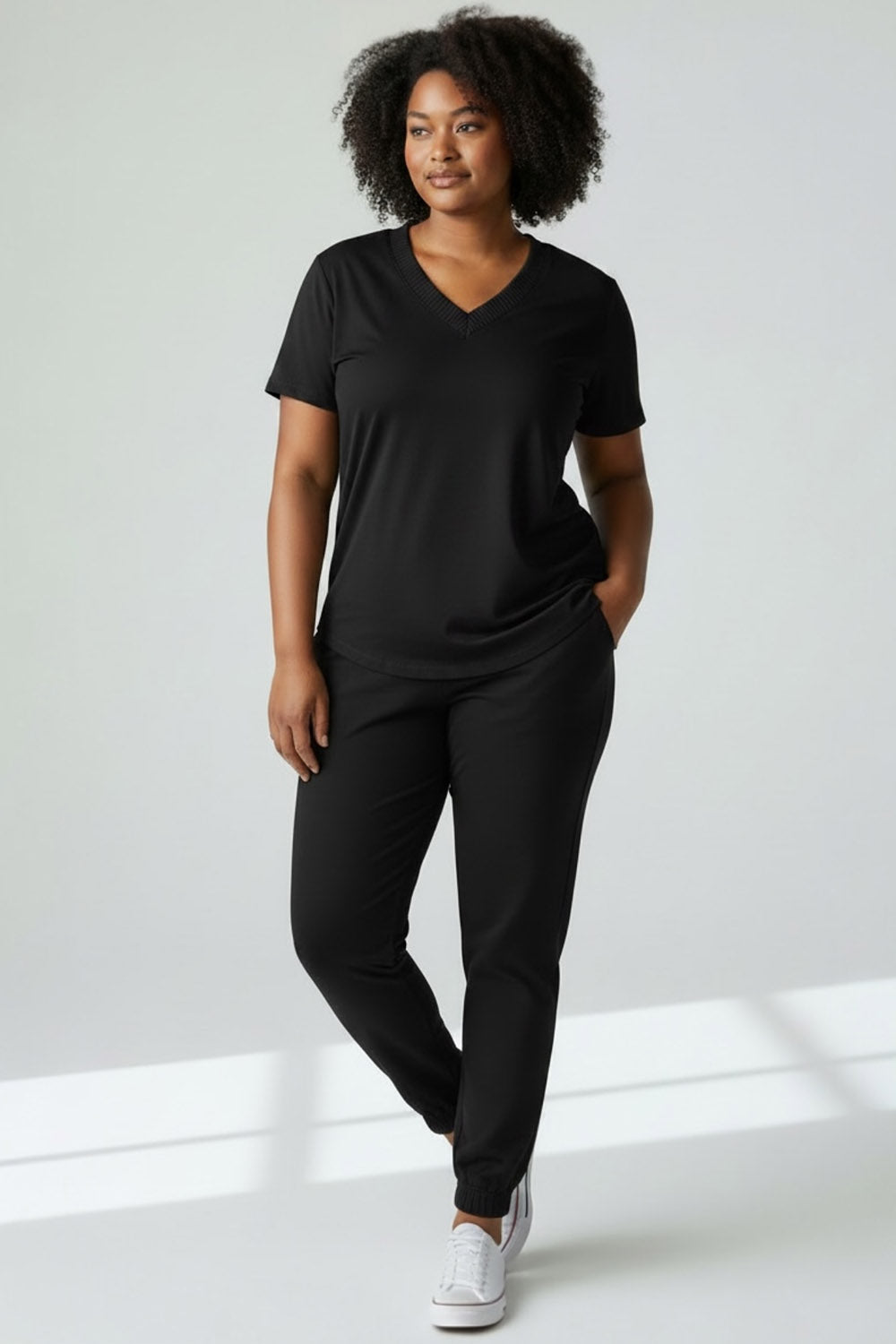A woman wearing the Advika V-neck t-shirt in Black with black jogging pants, standing in front of a white background. 