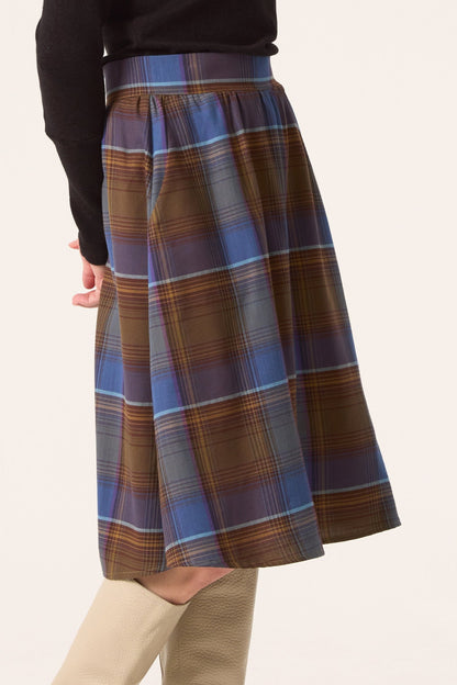 Waist-down view of a woman wearing the Alisma Skirt by Cokluch in Plaid Blue with a black turtleneck, standing in front of a white background 