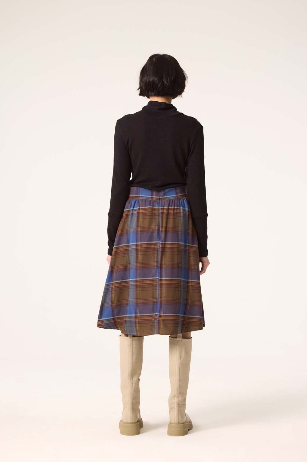 Back view of a woman wearing the Alisma Skirt by Cokluch in Plaid Blue with a black turtleneck, standing in front of a white background 