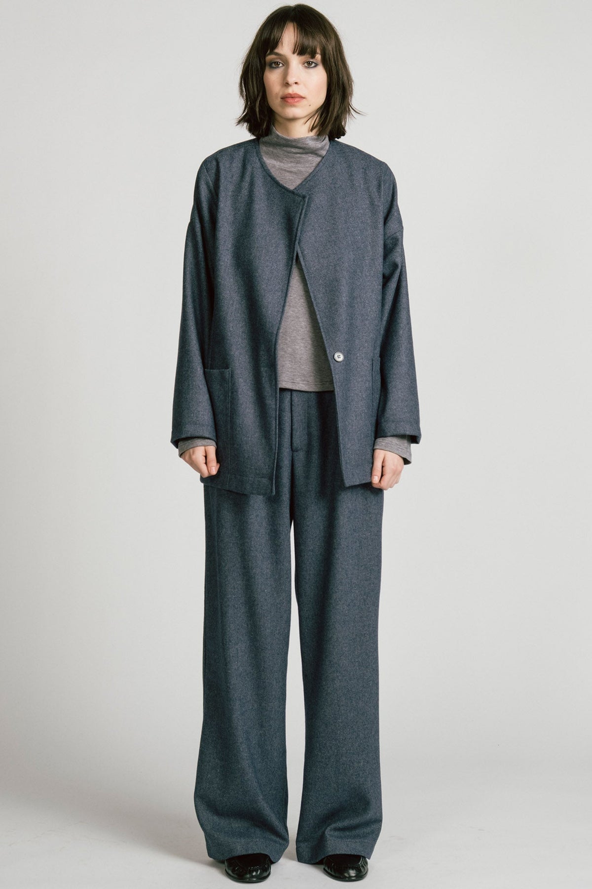 A woman wearing the Bootes Blazer by Allison Wonderland in Denim, a slouchy collarless blazer with a single waist button and a shoulder button, and drop sleeves, with large patch pockets. She is wearing it with the  Octans turtleneck in Grey and the matching Lynx Pants and standing against a light grey studio background.