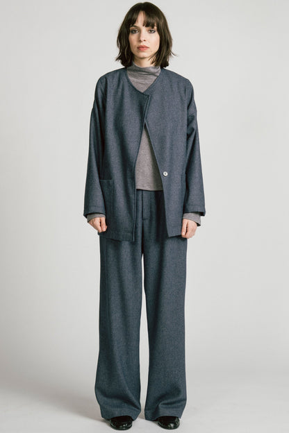 A woman wearing the Bootes Blazer by Allison Wonderland in Denim, a slouchy collarless blazer with a single waist button and a shoulder button, and drop sleeves, with large patch pockets. She is wearing it with the  Octans turtleneck in Grey and the matching Lynx Pants and standing against a light grey studio background.