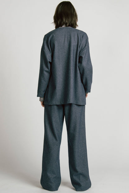 Back view of A woman wearing the Bootes Blazer by Allison Wonderland in Denim, a slouchy collarless blazer with a single waist button and a shoulder button, and drop sleeves, with large patch pockets. She is wearing it with the  Octans turtleneck in Grey and the matching Lynx Pants and standing against a light grey studio background.