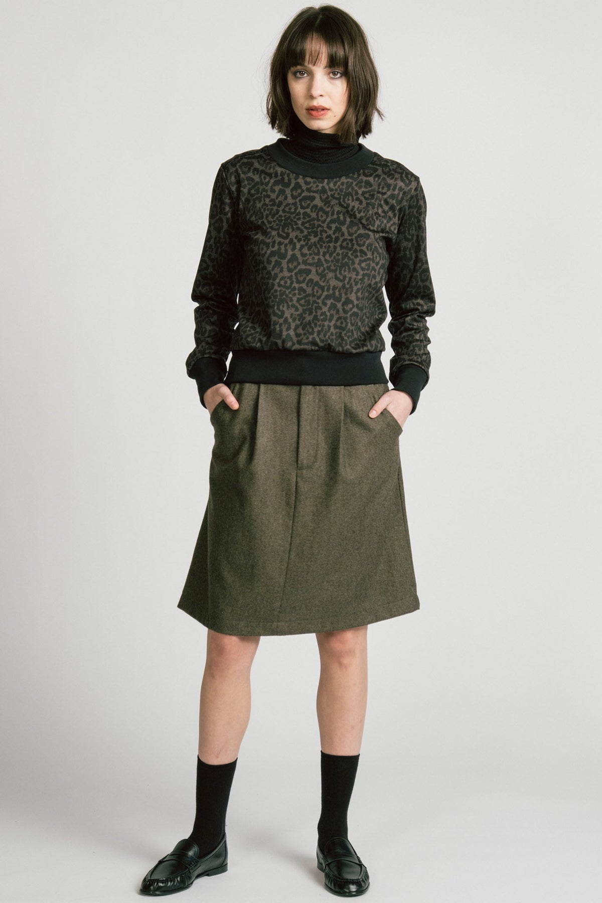 A woman wearing the Crux Sweatshirt by Allison Wonderland in Animal Print. she is wearing it with a grey skirt and standing in front of a white background. 