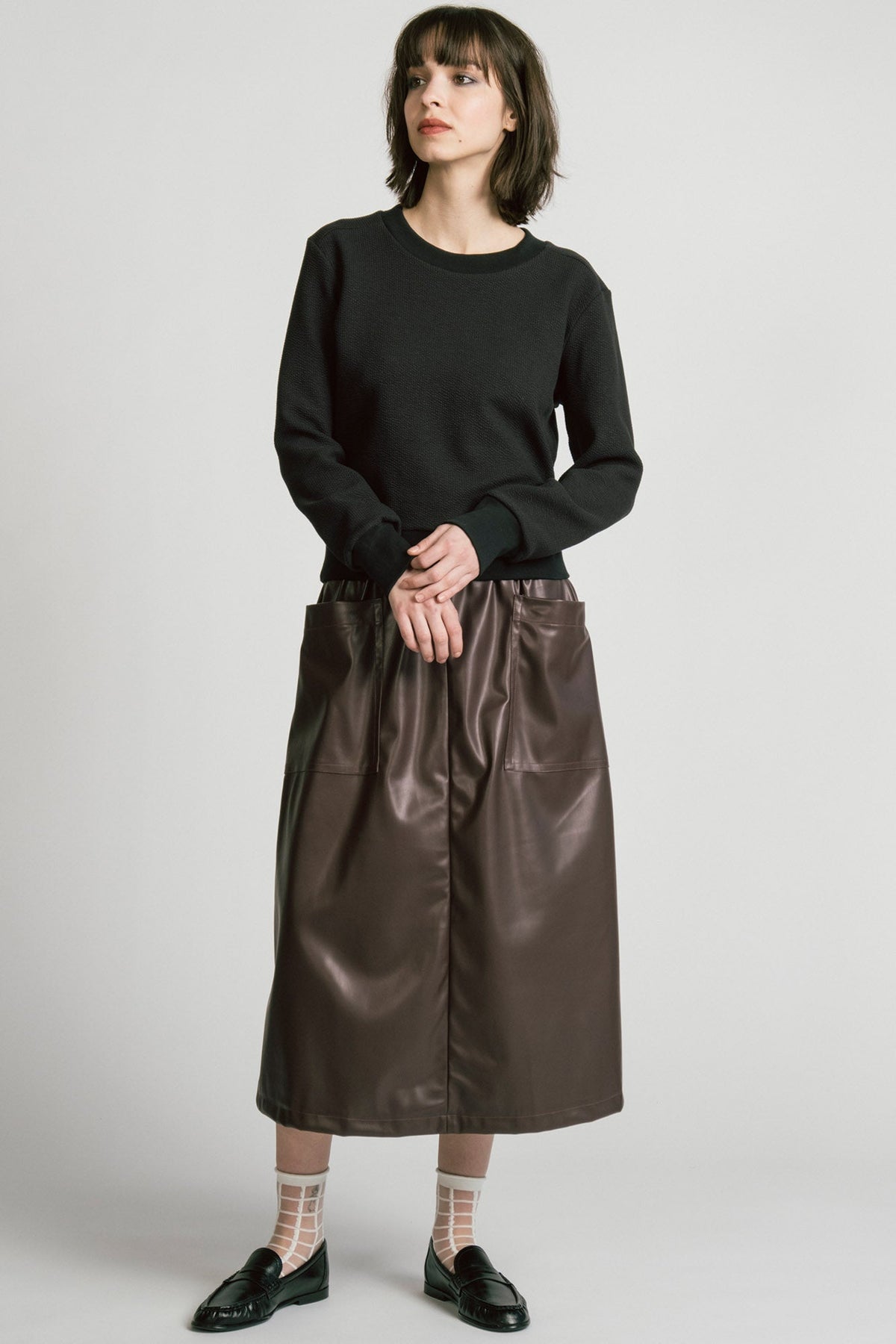 A woman wearing the Crux Sweatshirt by Allison Wonderland in Black Texture. she is wearing it with a brown pleather skirt and standing in front of a white background. 