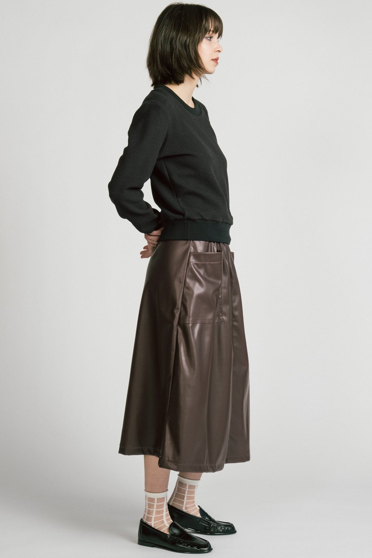 Side view of a woman wearing the Crux Sweatshirt by Allison Wonderland in Black Texture. she is wearing it with a brown pleather skirt and standing in front of a white background. 