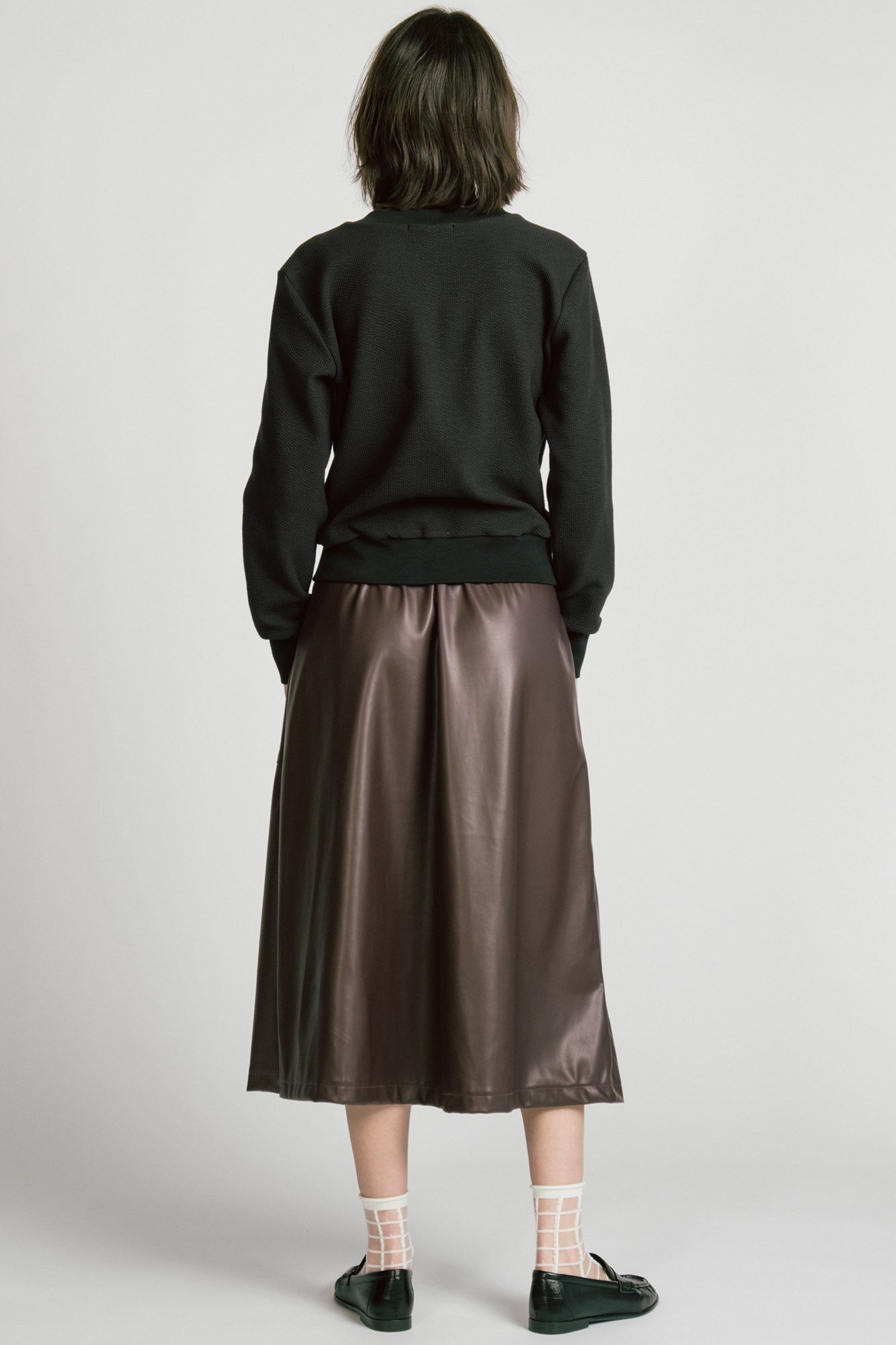 Back view of a woman wearing the Crux Sweatshirt by Allison Wonderland in Black Texture. she is wearing it with a brown pleather skirt and standing in front of a white background. 