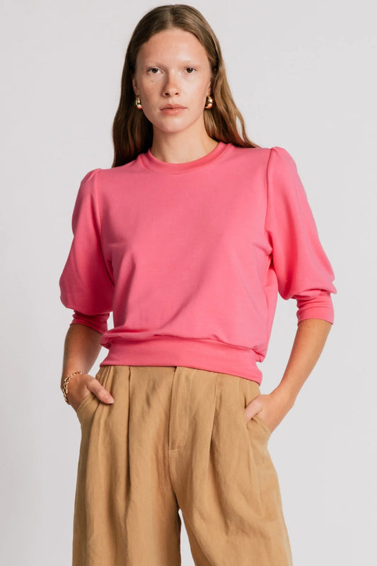 A woman wearing the Finch Sweatshirt by Allison Wonderland in Pink, a cropped sweatshirt with elbow-length puffed sleeves and ribbing at the neck and waistband. She is wearing it with beige pants and standing in front of a white background.