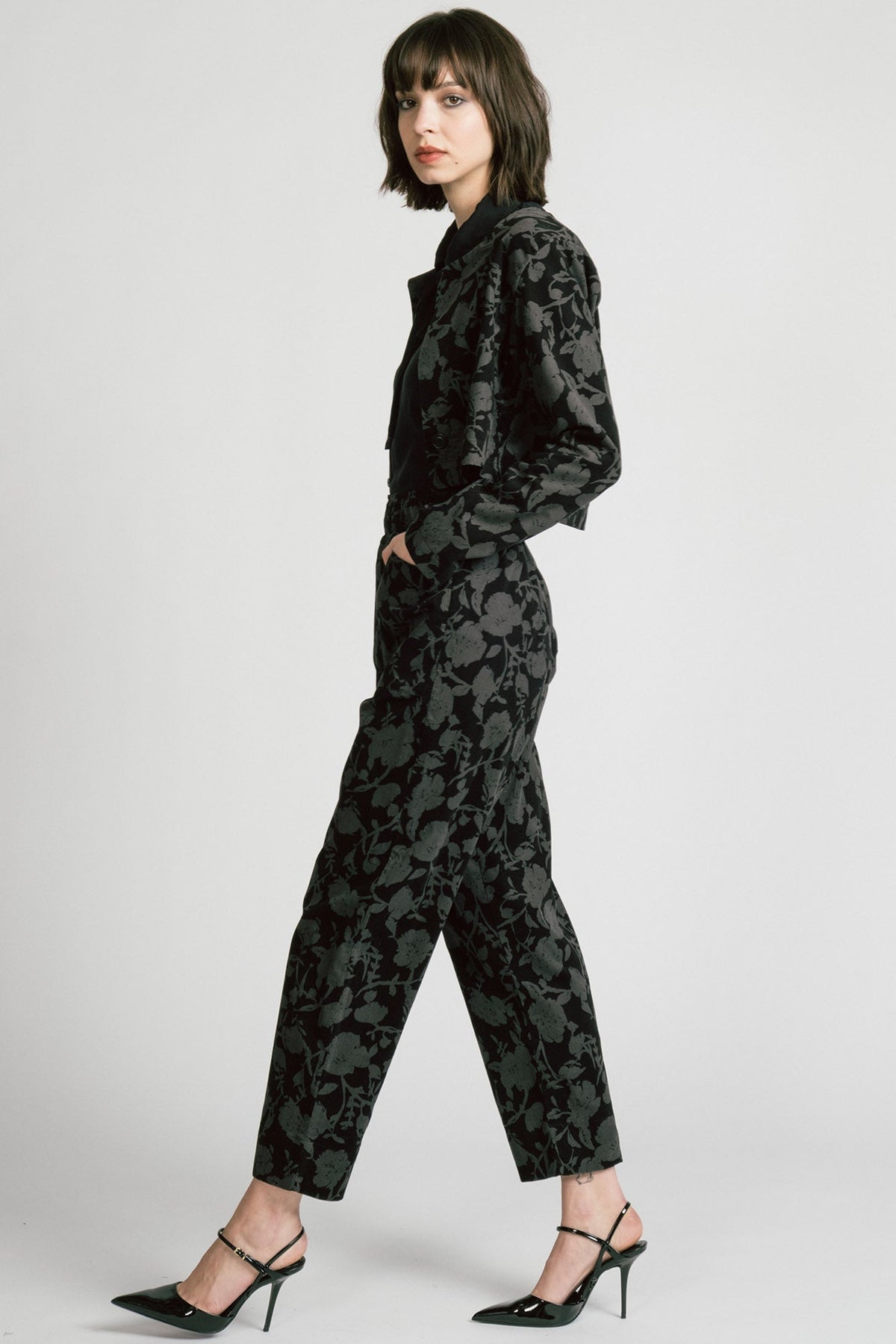 Side view of a woman wearing the Libra Bolero by Allison Wonderland in Floral, a dark printed corduroy cropped jacket with a single button closure. She is wearing it over a black top with matching pants. 