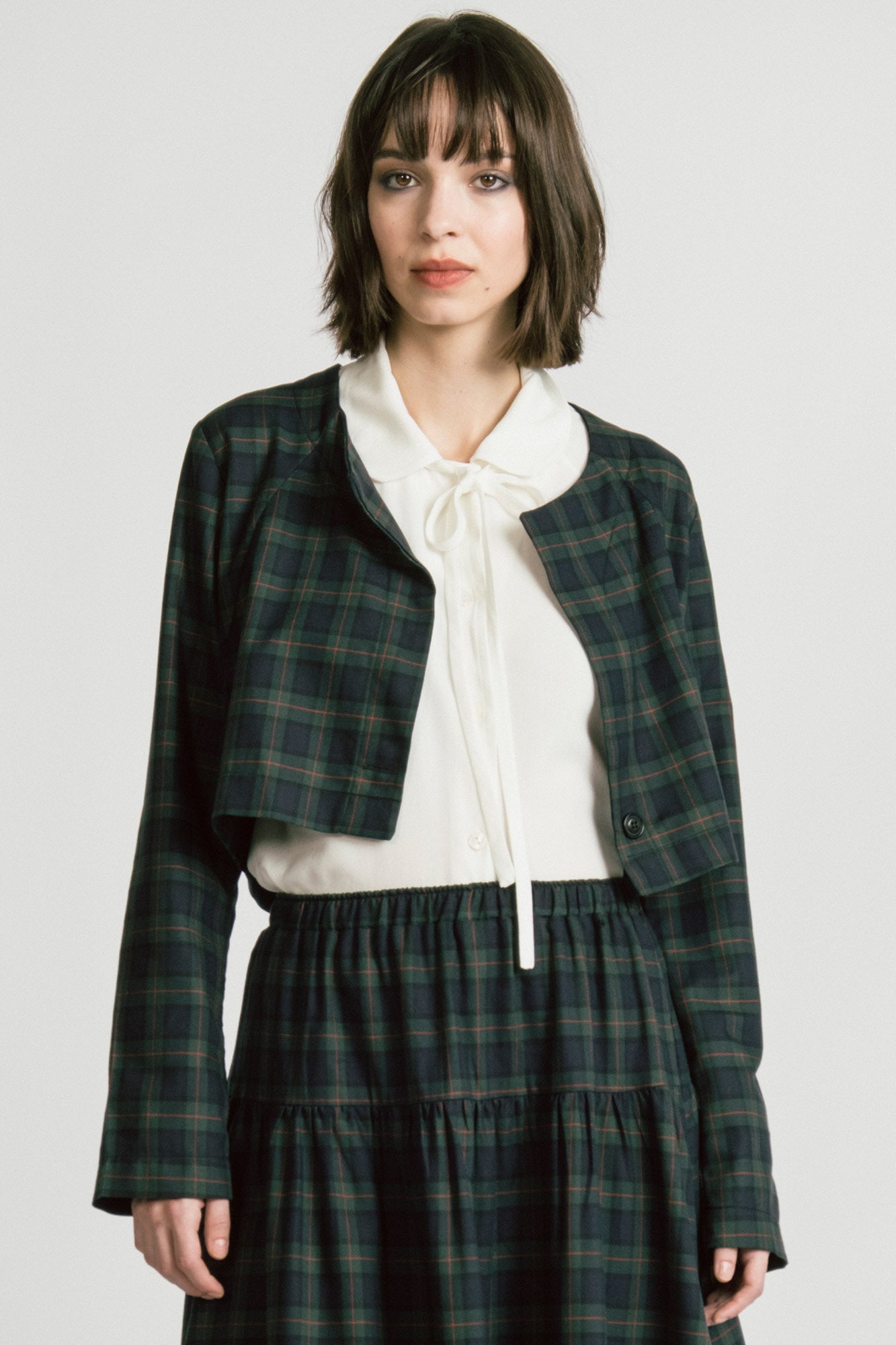 A woman wearing the Libra Bolero by Allison Wonderland in Plaid, a green plaid cropped jacket with a single button closure. She is wearing it over a white top with a matching skirt. 