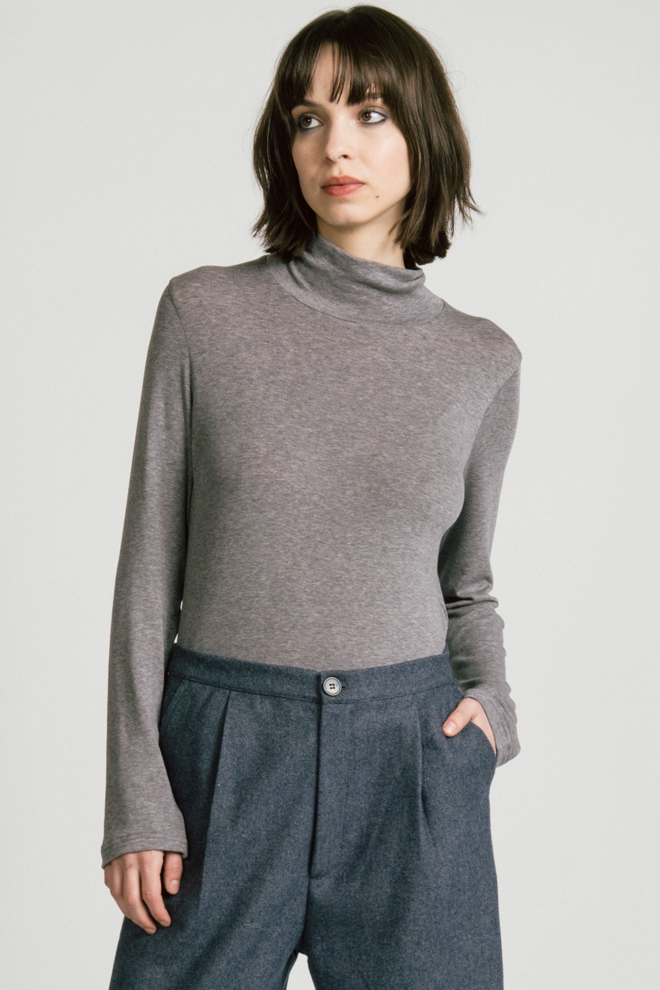 A woman wearing the Octans Turtleneck by Allison Wonderland in Heather Grey, with dark grey pleated trousers, standing against a white studio background.