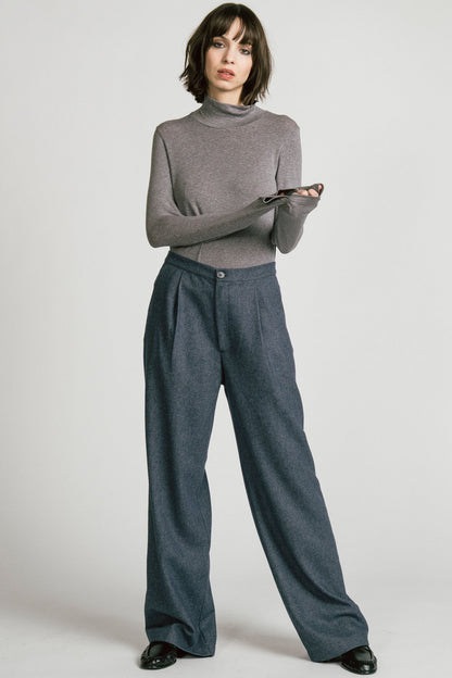 A woman wearing the Octans Turtleneck by Allison Wonderland in Heather Grey, with dark grey pleated trousers, standing against a white studio background.