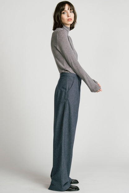 Side view of a woman wearing the Octans Turtleneck by Allison Wonderland in Heather Grey, with dark grey pleated trousers, standing against a white studio background.