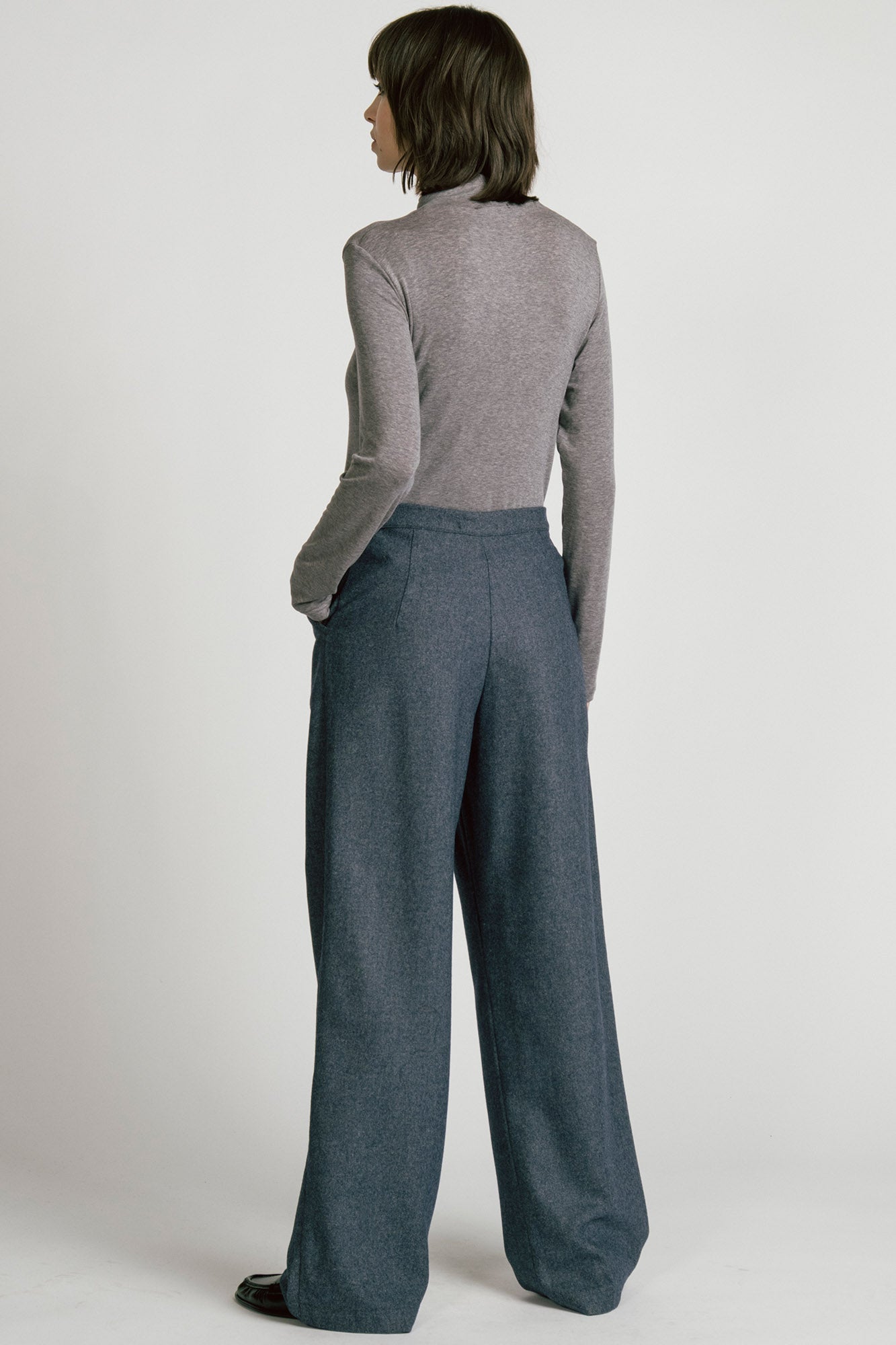 Back view of a woman wearing the Octans Turtleneck by Allison Wonderland in Heather Grey, with dark grey pleated trousers, standing against a white studio background.