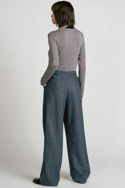 Back view of a woman wearing the Octans Turtleneck by Allison Wonderland in Heather Grey, with dark grey pleated trousers, standing against a white studio background.