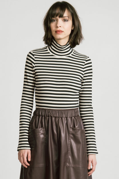 A woman wearing the Octans Turtleneck by Allison Wonderland in black and white Stripe, a ribbed turtleneck. She is wearing it with a brown pleather skirt and standing against a white studio background.