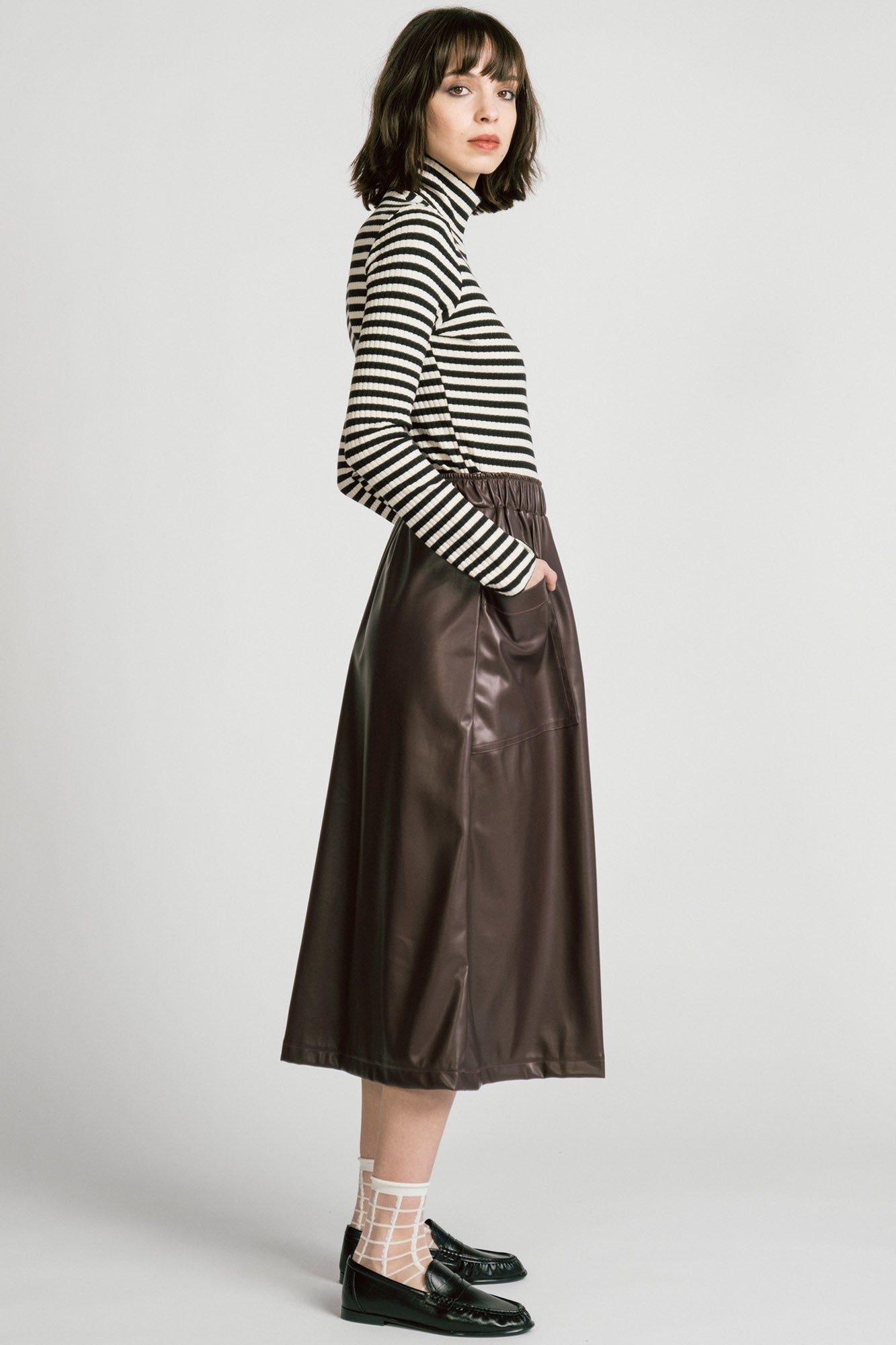 Side view of a woman wearing the Octans Turtleneck by Allison Wonderland in black and white Stripe, a ribbed turtleneck. She is wearing it with a brown pleather skirt and standing against a white studio background.