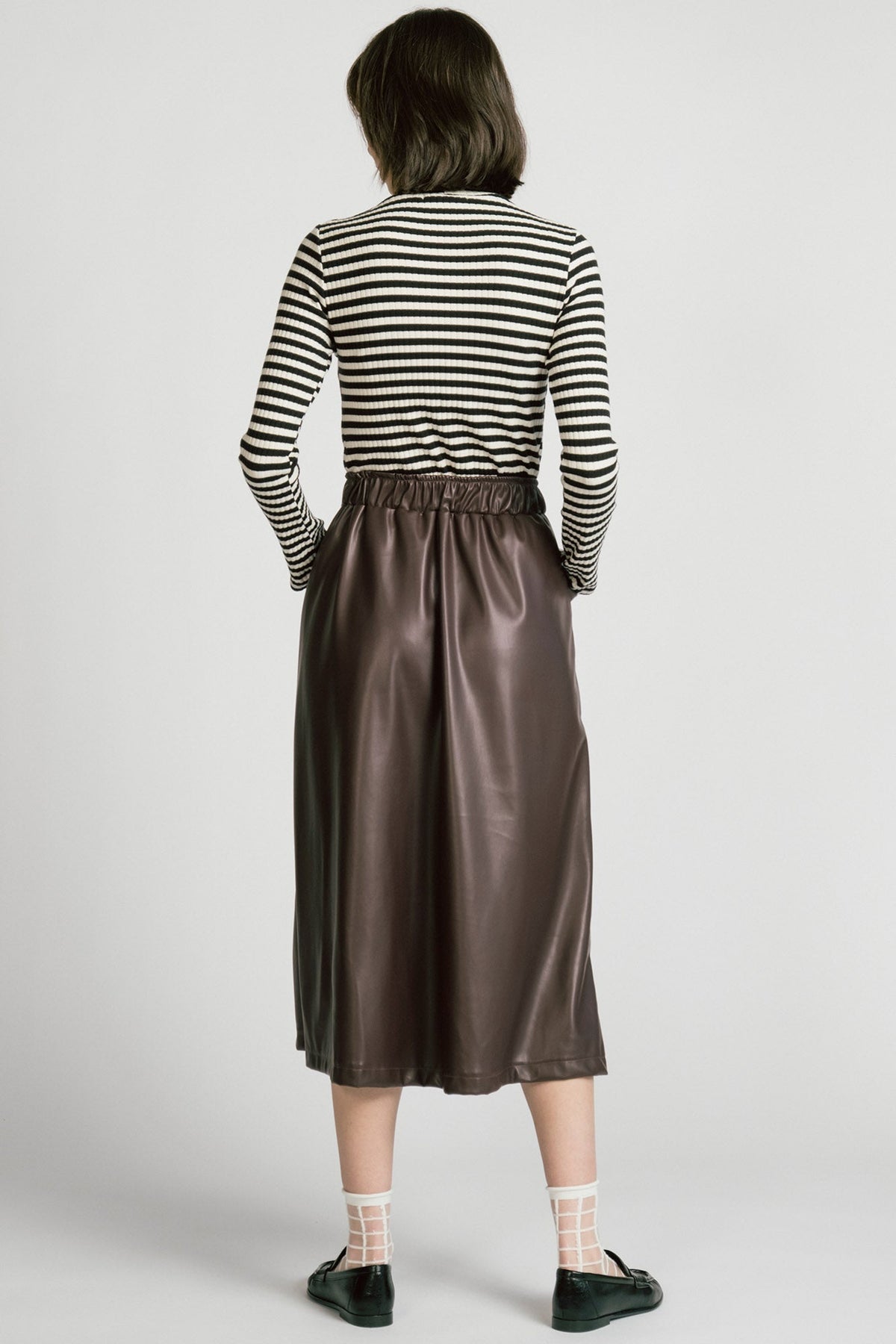 Back view of a woman wearing the Octans Turtleneck by Allison Wonderland in black and white Stripe, a ribbed turtleneck. She is wearing it with a brown pleather skirt and standing against a white studio background.