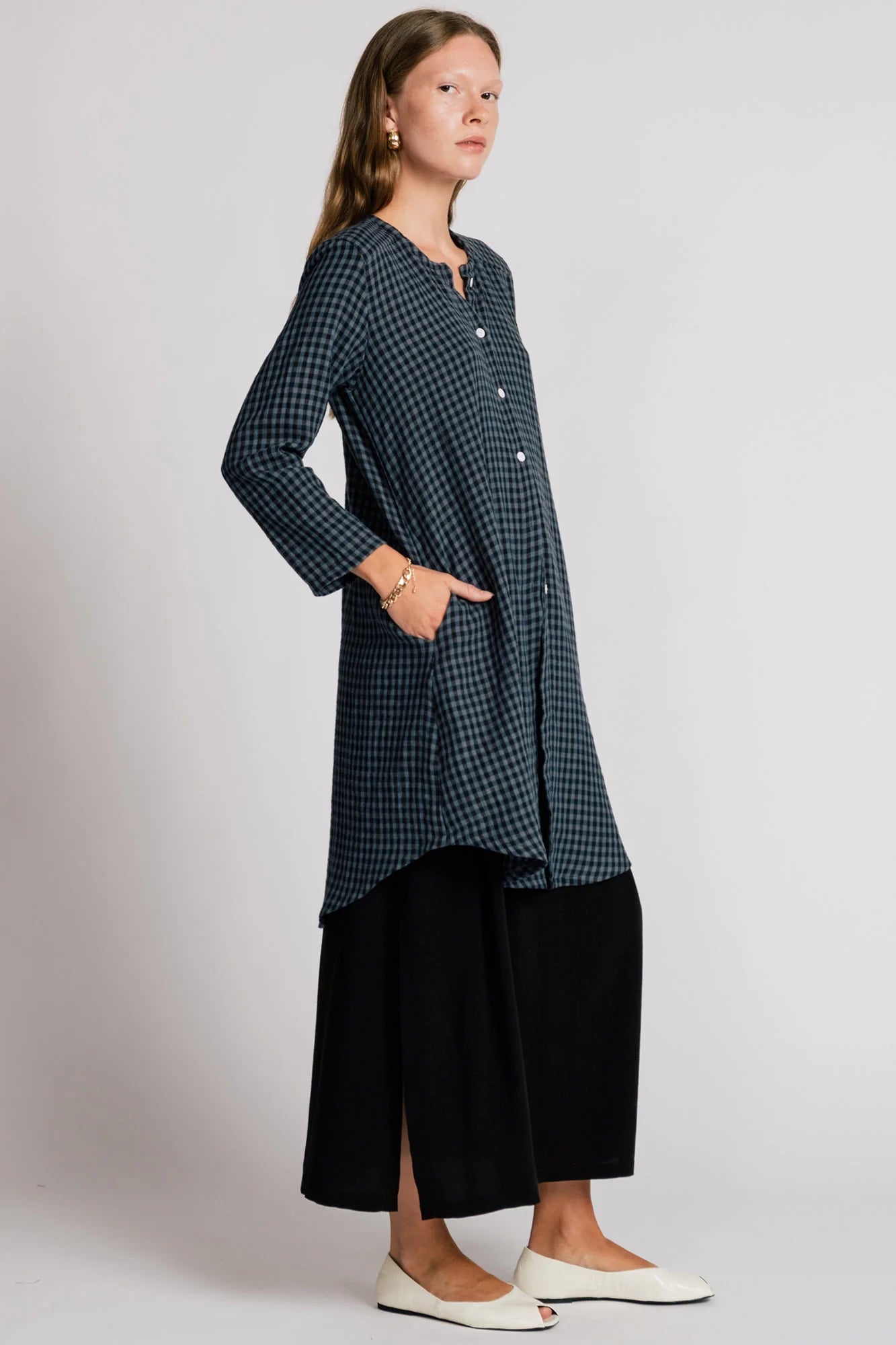 Side view of a woman wearing the Oriole Tunic by Allison Wonderland in Navy Gingham, a long tunic with a button front and rounded hi-low hemline. She is wearing it over a black skirt and standing against a grey background. 